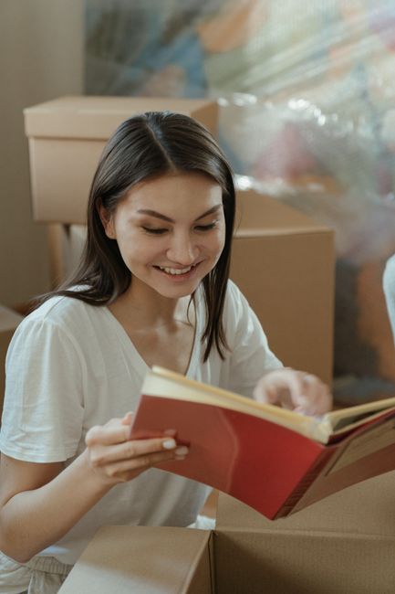 Inside a cardboard moving box viewed from above, containing a black and white photo album with the title 'HomeBook' on the cover, which displays multiple images of living room interior scenes such as 