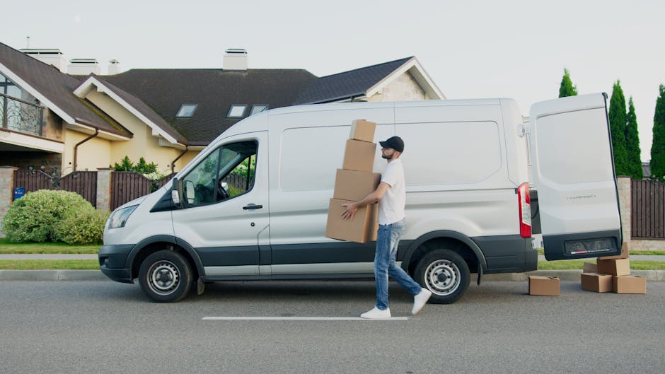 In a driveway or loading area during the daytime, two people are engaged in a home relocation process involving furniture and packing materials. A man wearing a white polo shirt, a black cap, and wire