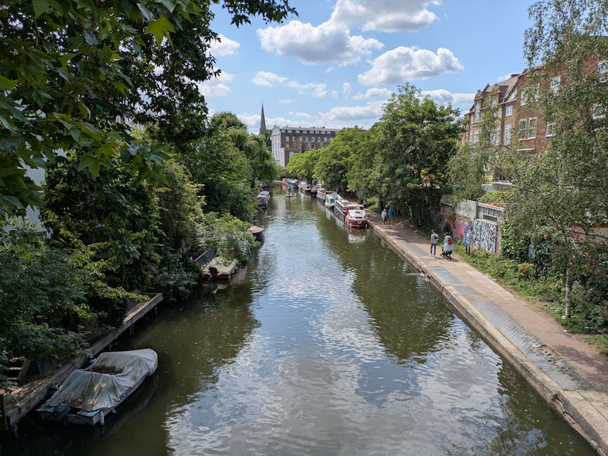 A view of a narrow canal lined with lush green trees and residential buildings on the right side along a paved walkway in Merton. Several boats are docked along the canal, some covered with protective