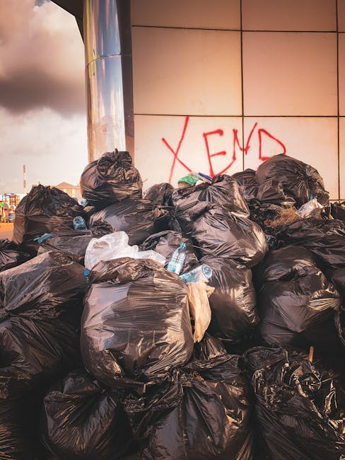 A row of four large white rubbish bins with fitted lids, positioned on a paved surface next to a wooden barrier fence. To the left of the bins, there is a smaller dark-colored bin labeled 'PAPER' and 