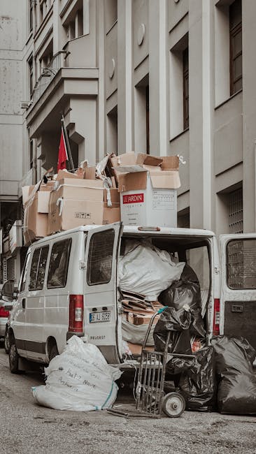A white moving van parked on a city street next to a multi-storey building with large windows and balconies. The van’s rear doors are open, revealing various packing materials, including cardboard b